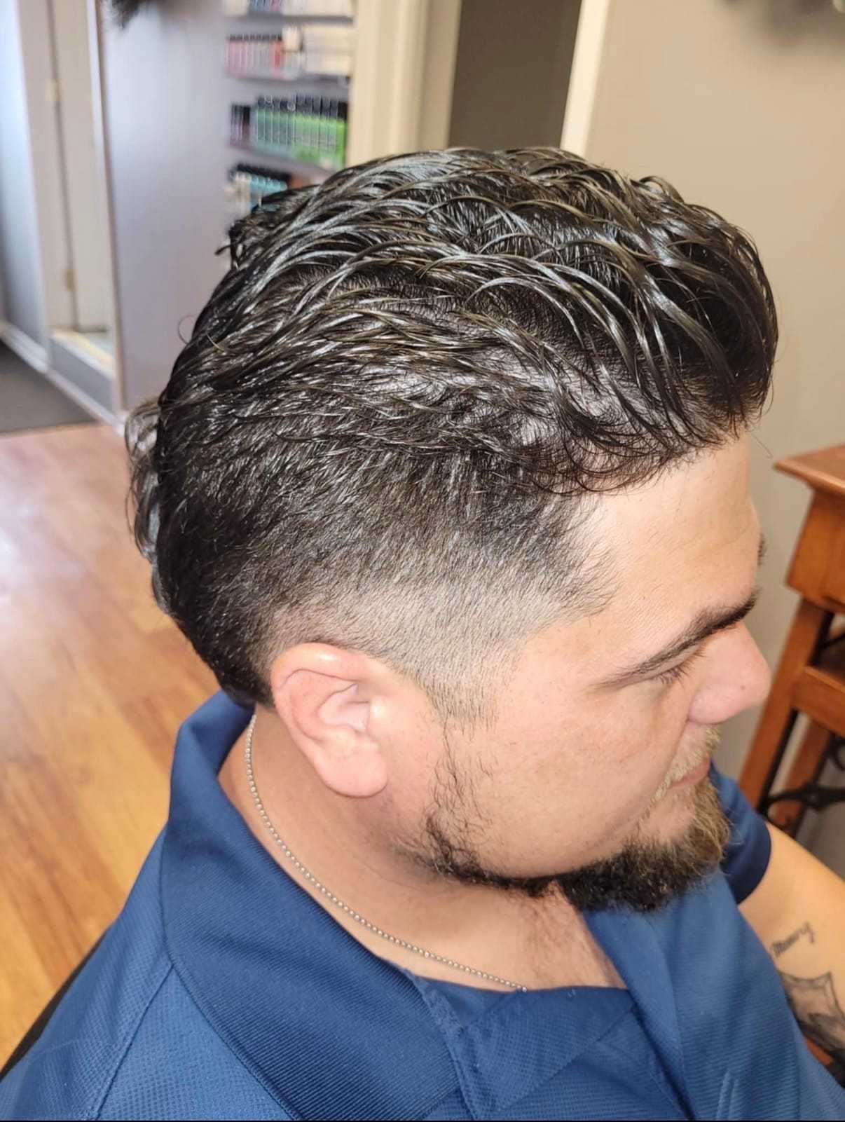 Man with styled hair and beard in a blue shirt, sitting indoors on a wooden floor.