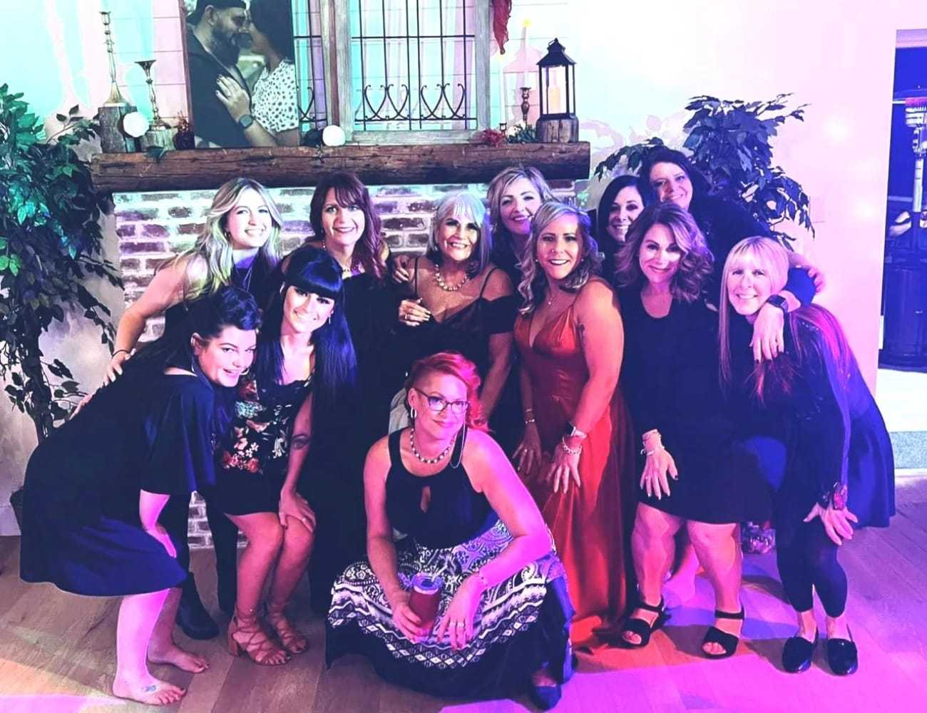 Group of women posing together indoors, smiling happily near a fireplace.